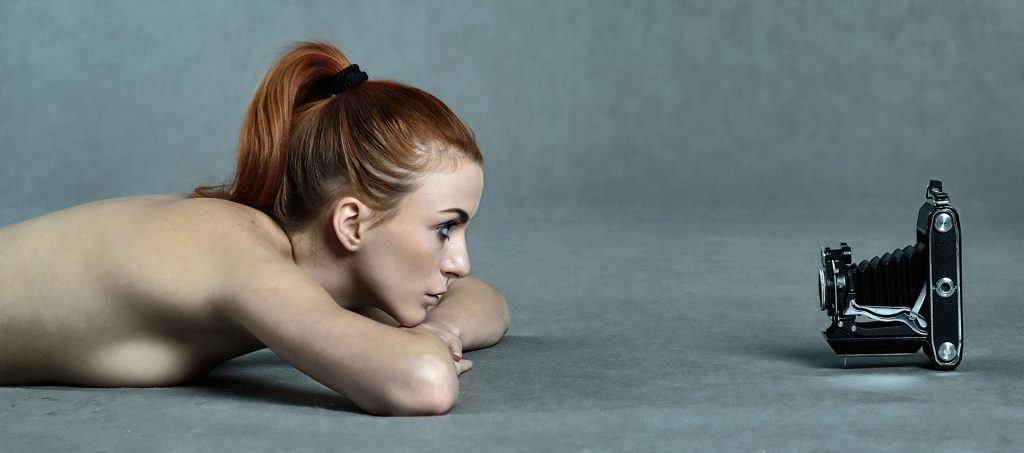 A bare-shouldered woman with red hair lies on a gray floor, looking intently at a vintage bellows camera