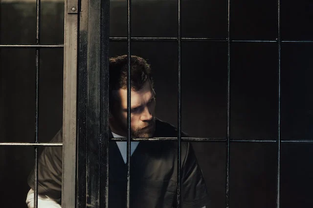 A man in prison that's leaning against the bars of his cell.