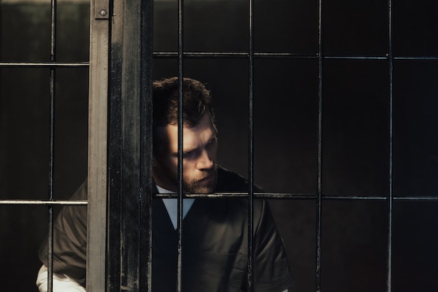 A man in prison that's leaning against the bars of his cell.